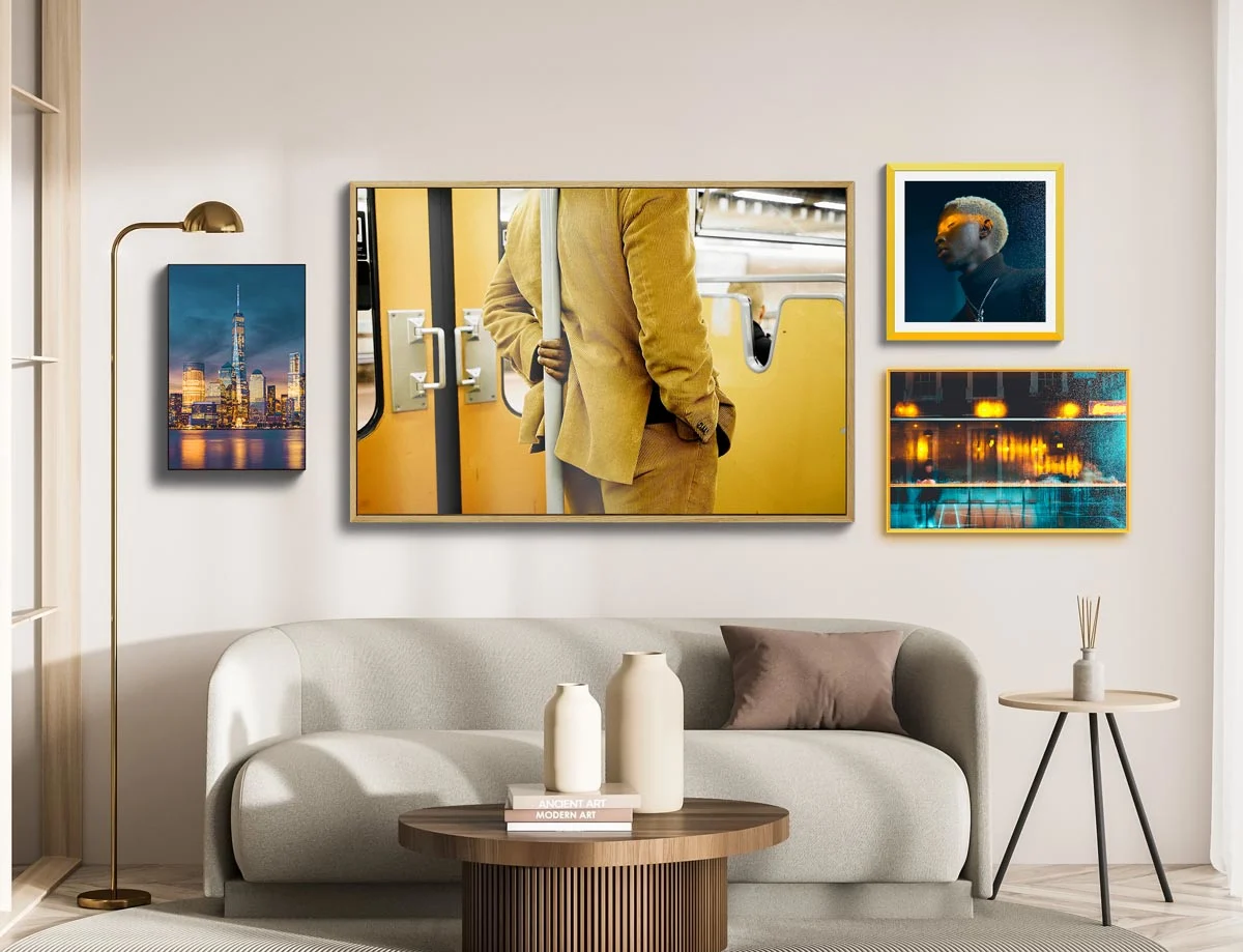 A living room with a beige sofa, a round wooden table and various framed pictures on the wall. A golden floor lamp stands to the left of the sofa.