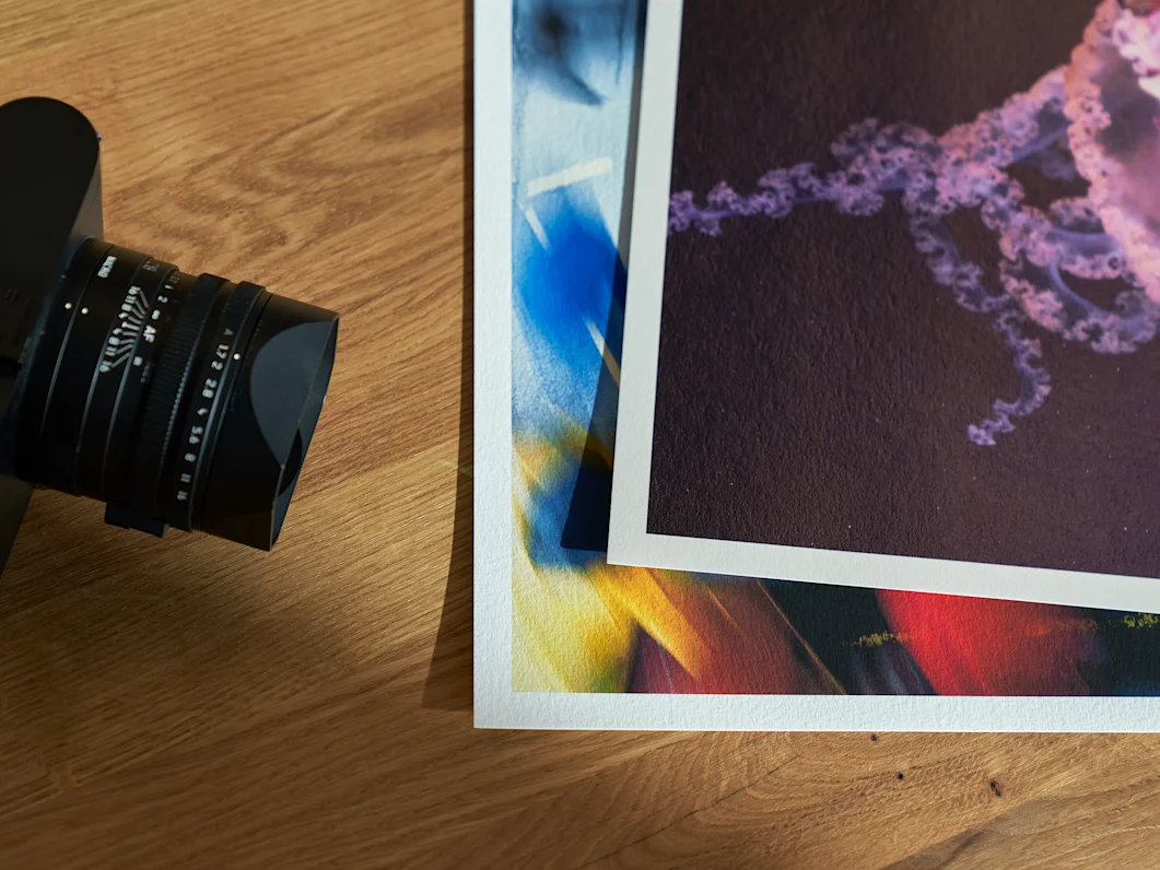 Fine Art Print 3 Two Fine Art paper prints with abstract colorful patterns lie on a wooden table next to a camera lens.