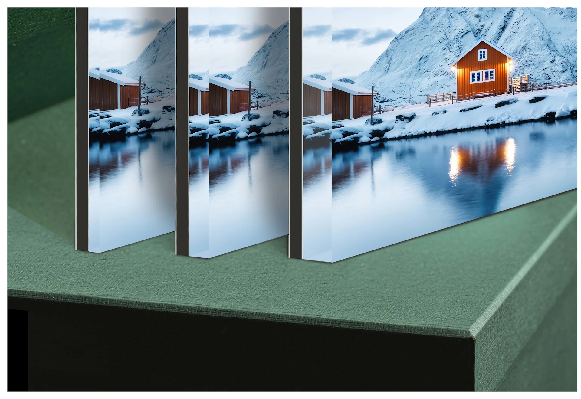 Three pictures of different thicknesses behind acrylic glass are placed on a green gift box. The image shows a snowy landscape with an orange wooden house.