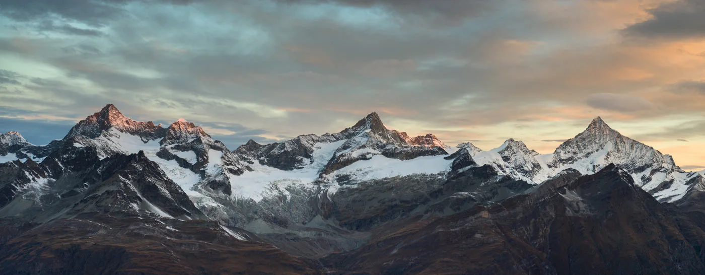 Mountain range with snow-covered peaks under a cloudy sky at sunset.