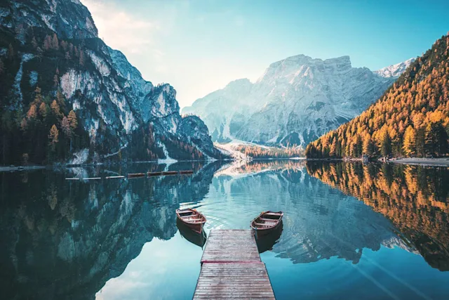 Wooden jetty with two rowboats on a tranquil mountain lake, surrounded by wooded and rocky mountains in autumn.
