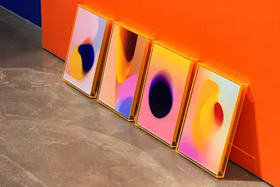 Four colorful abstract paintings in orange frames lean against an orange wall on a gray floor.