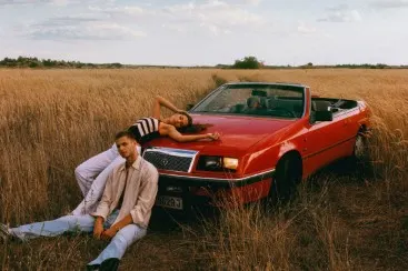 Two people in a red convertible parked in a field of high, dry grass, sky with clouds.