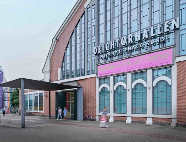 Die Fotografie zeigt die markante Backsteinfassade der Deichtorhallen mit ihren großen, bogenförmigen Glasfenstern. Über dem Eingangsbereich prangt der große Schriftzug des Gebäudes, darunter hängt ein langes, auffallend pinkes Ausstellungsbanner. Auf dem gepflasterten Vorplatz im Vordergrund sind vereinzelte Passanten in Bewegung sowie Fahrräder an den Wänden zu erkennen. Die weitwinklige Außenaufnahme fängt die urbane und kulturelle Atmosphäre des Ausstellungsortes an einem leicht bewölkten Tag ein.