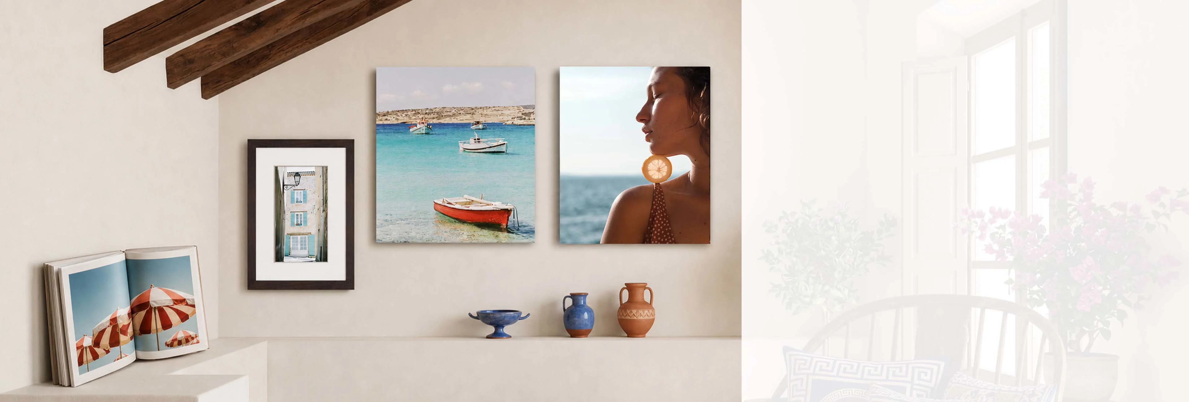 Este banner de formato de ancho extremo muestra una elegante presentación de pared con varias obras de arte en una habitación luminosa de estilo mediterráneo. En el centro cuelgan dos impresiones cuadradas sin marco: una muestra barcos en un mar turquesa y la otra el perfil de una mujer con el océano de fondo. A la izquierda, una foto vertical enmarcada y un libro de arte abierto apoyado en un saliente completan la composición, realzada por jarrones de cerámica antigua debajo. La mitad derecha de la imagen se desvanece suavemente hacia una habitación luminosa y soleada con una silla de madera y flores rosas.