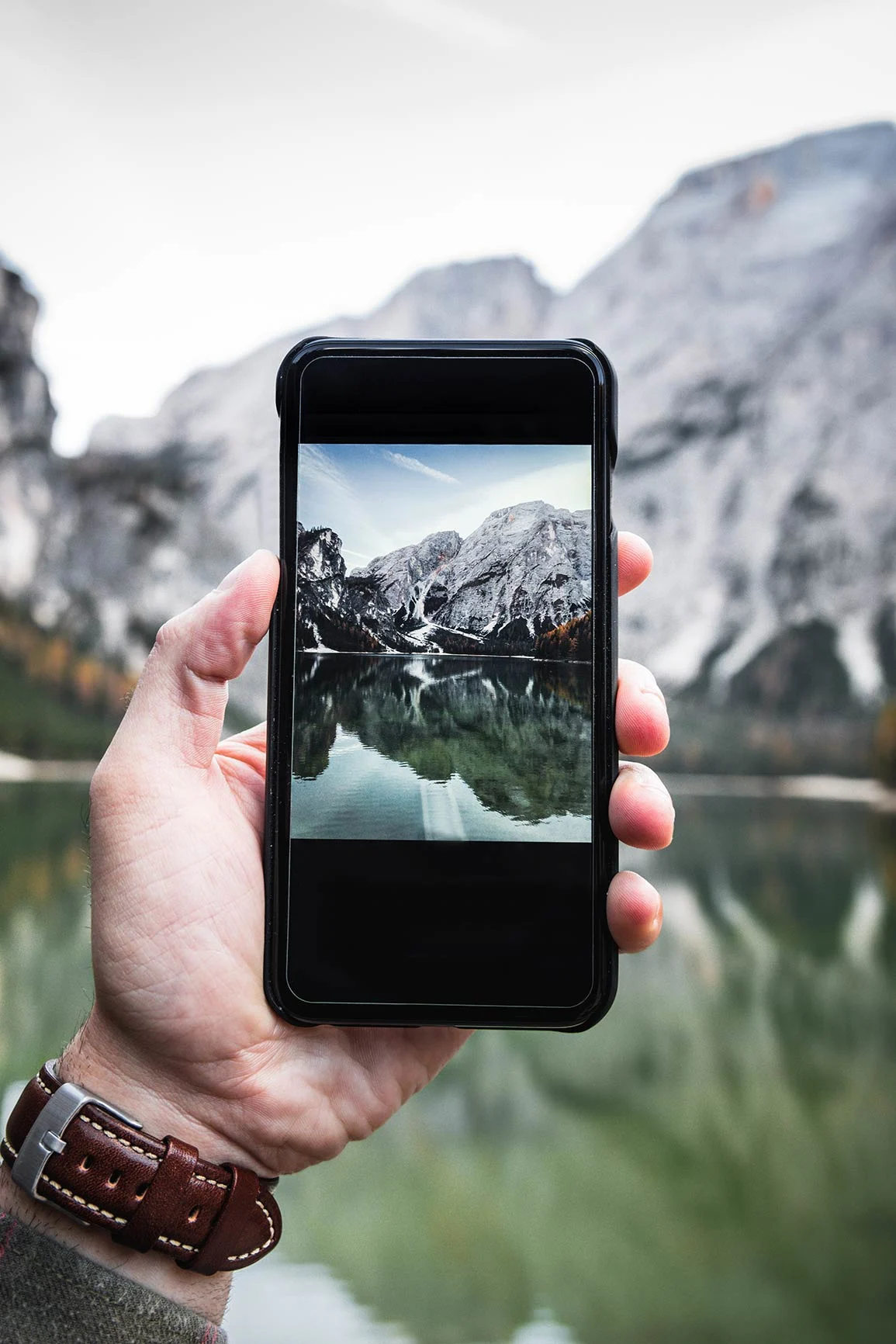 Hand holding smartphone with mountain lake motif, blurred mountain lake with rocks and water in the background.