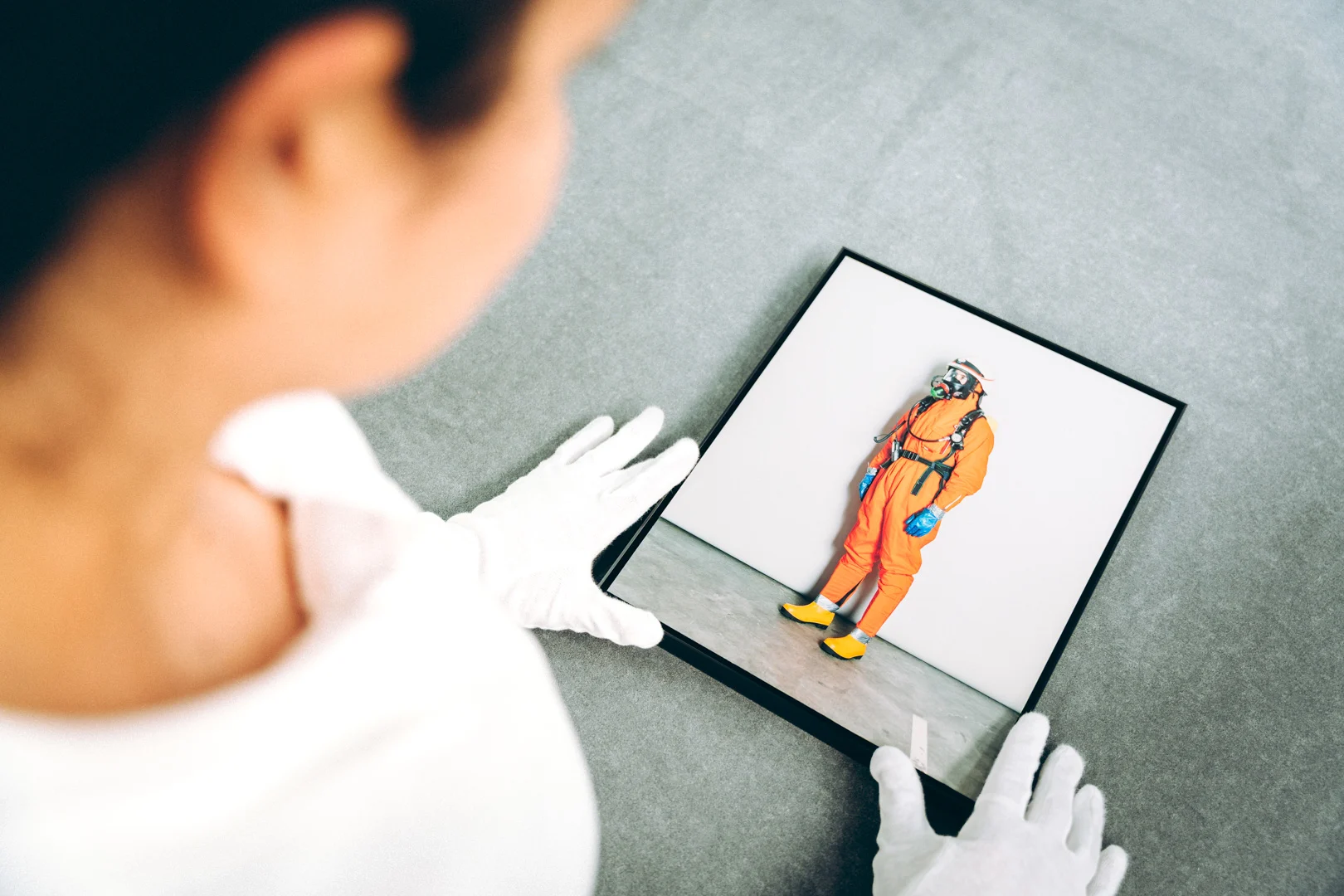 A woman seen from behind, wearing white gloves and a white shirt, gazing at a framed artwork.