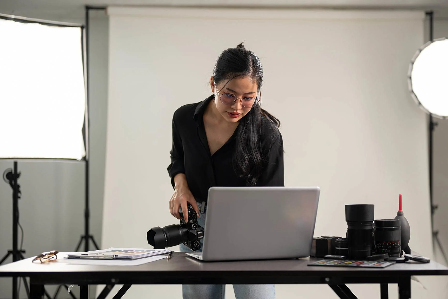A woman is standing at a table with a laptop, a camera, and camera lenses in front of two large photography lights.