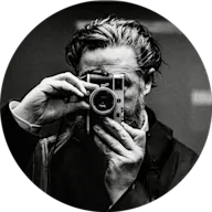 Ein bärtiger Fotograf in dunkler Kleidung macht ein Foto mit einer klassischen Messsucherkamera im kreisförmigen Schwarzweiß-Porträtformat