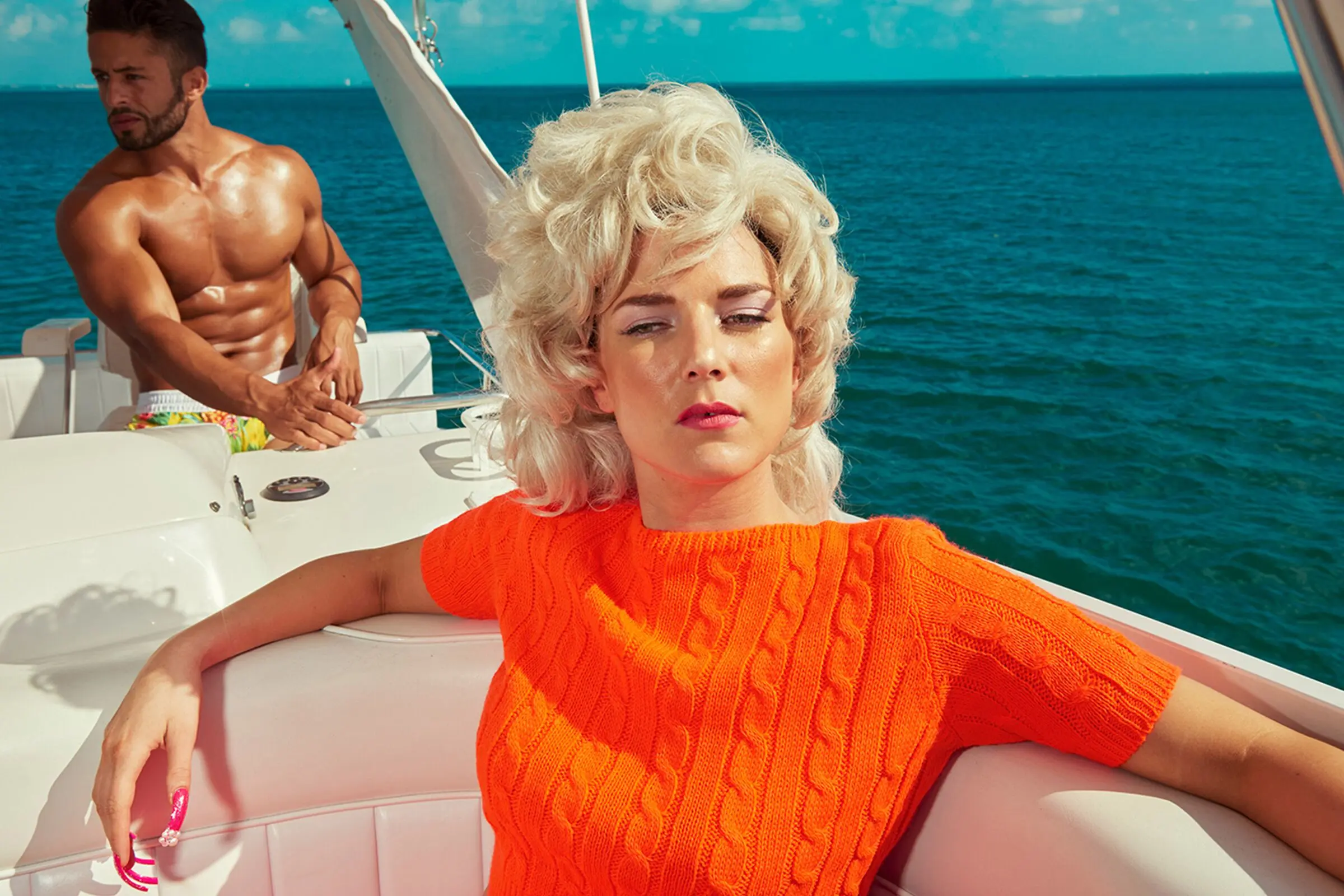 Kourtney Roy self portrait wearing bright orange shirt on a boat with tanned muscular driver in the background.