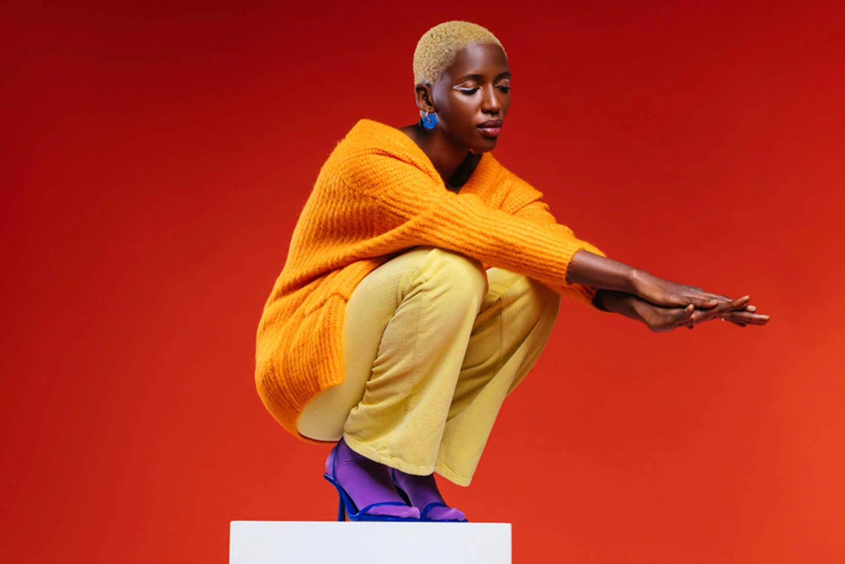 A woman with short blonde hair is sitting on a white platform against a red background; she is wearing an orange sweater, yellow pants, and blue shoes.