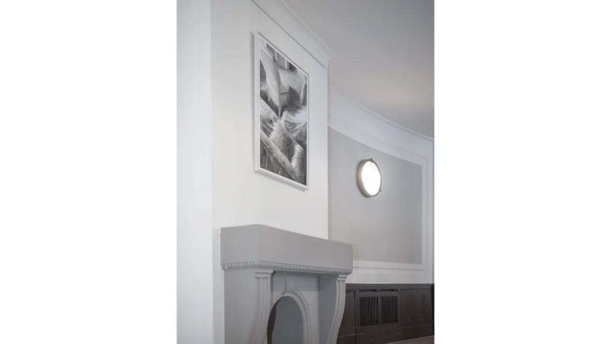 A white ornate fireplace mantel below a framed black and white abstract artwork, with a round wall light on a grey wall.