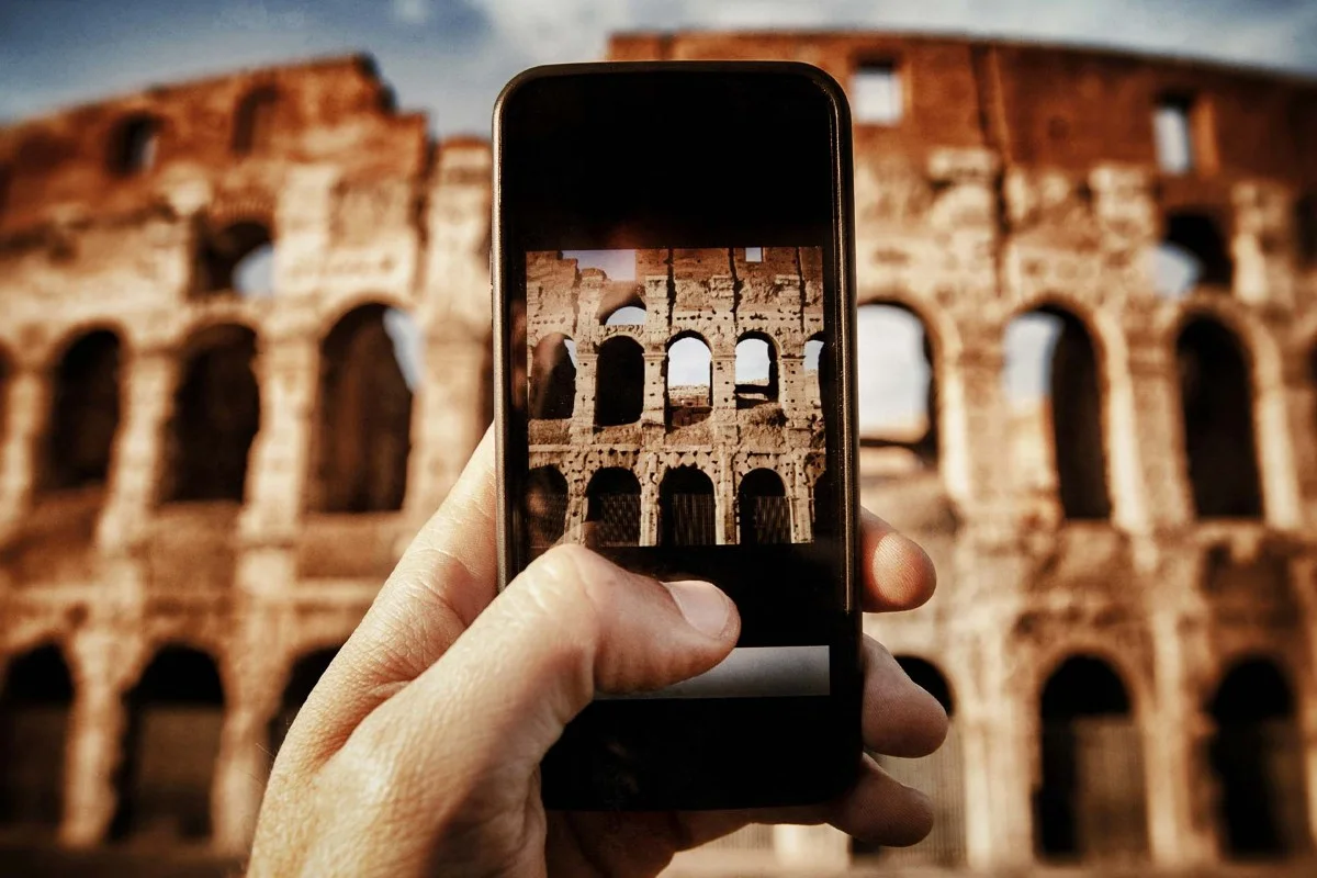 Una mano sostiene un smartphone que muestra una foto del Coliseo, con las ruinas romanas desenfocadas al fondo.