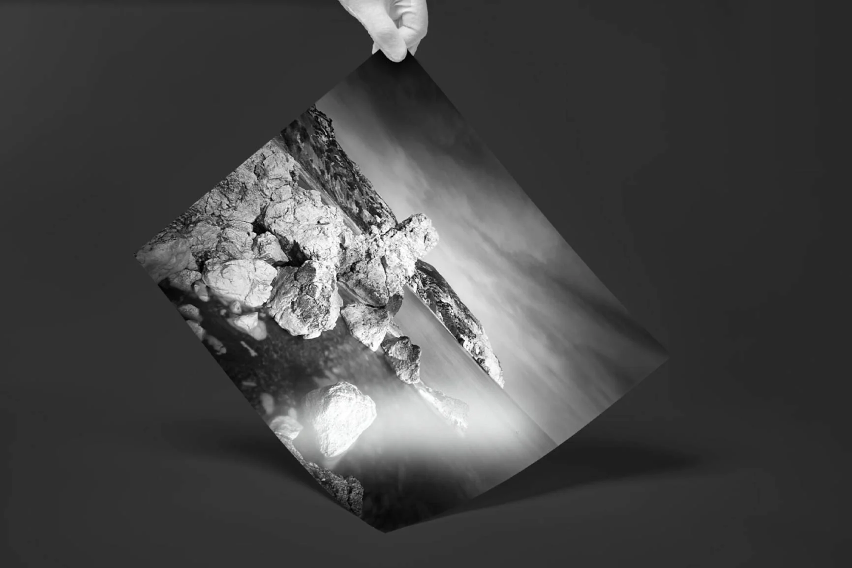 Mano che regge una fotografia in bianco e nero con rocce e un arco naturale sul mare.