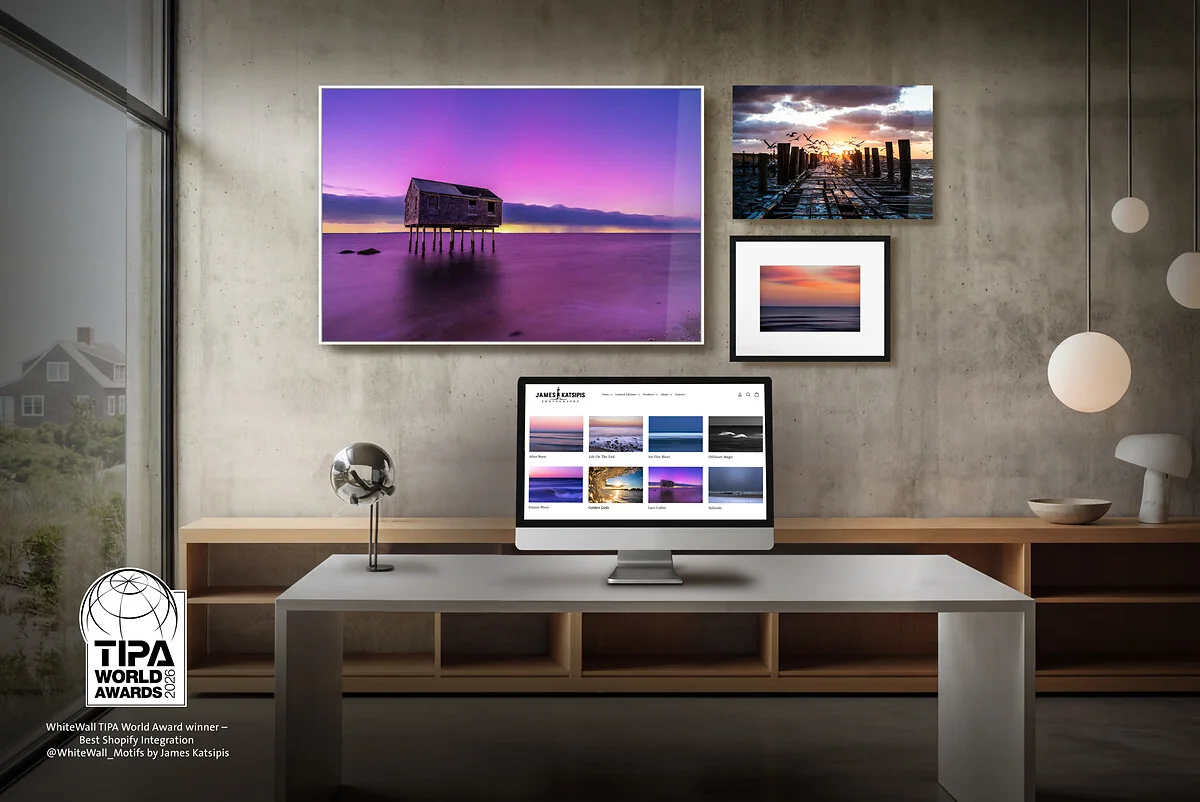 Ein modernes Arbeitszimmer mit Sichtbetonwand, auf dem ein Desktop-Monitor eine Fotografie-Webseite zeigt. Darüber hängen drei Landschaftsaufnahmen: ein großes purpurfarbenes Küstenmotiv im weißen Rahmen sowie zwei kleinere Bilder eines Stegs bei Sonnenuntergang. Unten links ist das Logo der „TIPA World Awards 2026“ für die beste Shopify-Integration zu sehen.