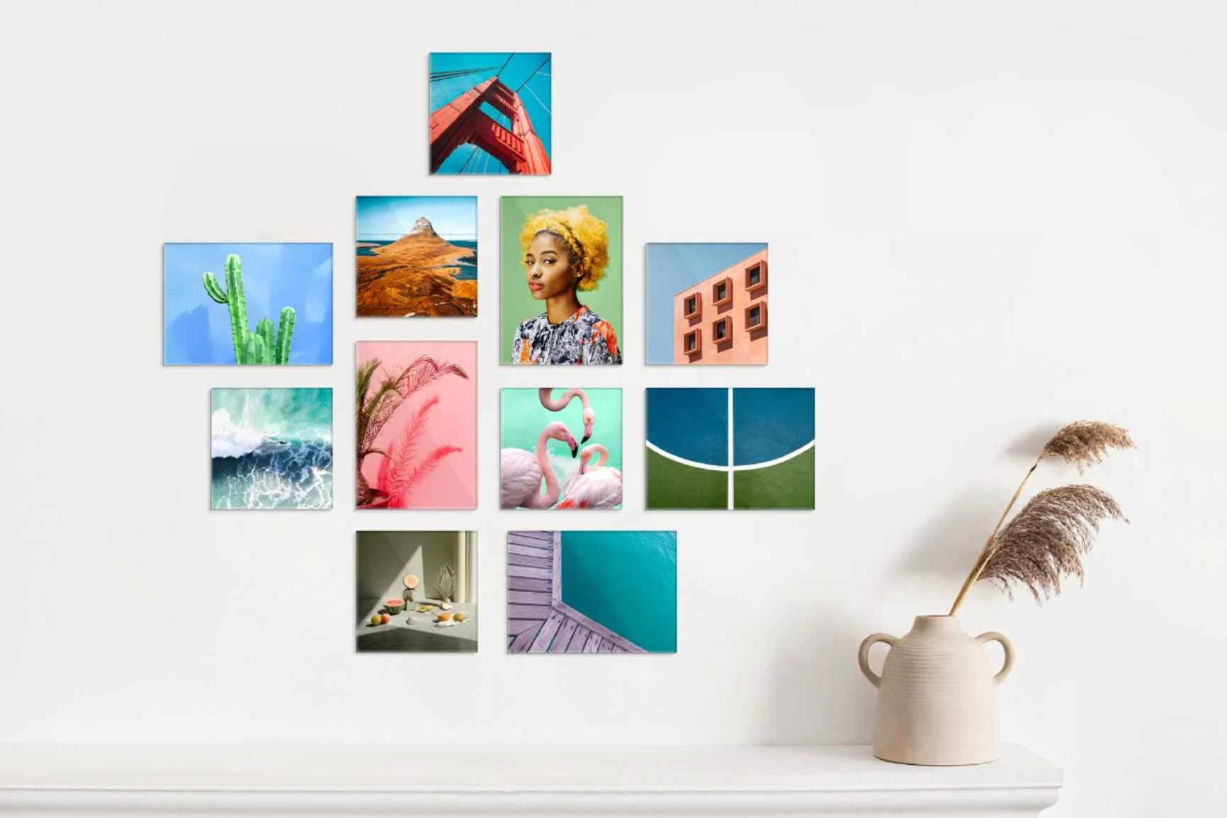 Parete con una composizione di diverse foto colorate in vari formati dietro del plexiglass sopra una mensola bianca con un vaso e canne di giunco.