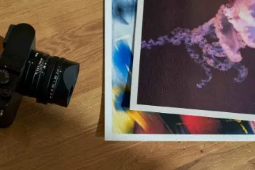 Zwei Fine Art Prints mit abstrakten, farbigen Mustern liegen auf einem Holztisch neben einem Kameraobjektiv.