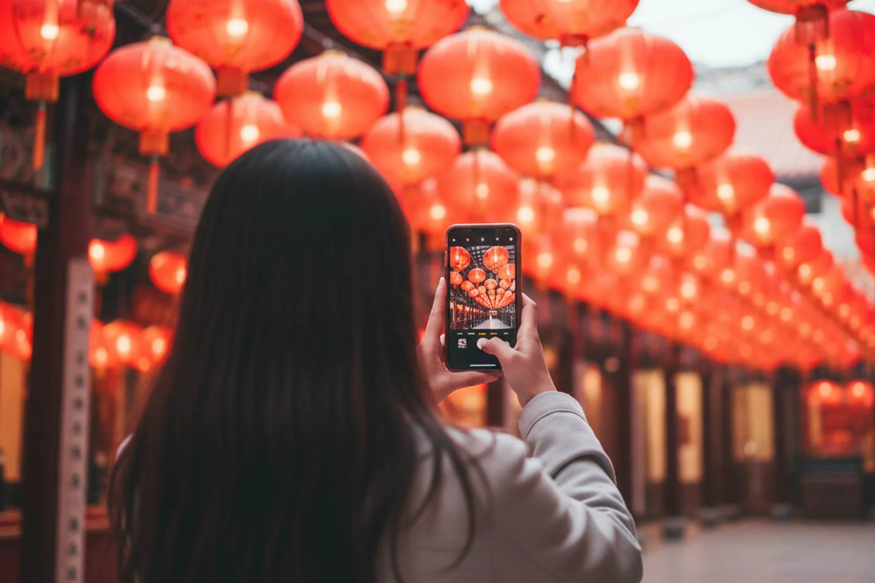 Frau mit langen dunklen Haaren fotografiert mit ihrem Smartphone eine Decke aus vielen roten Laternen in einem Gang.