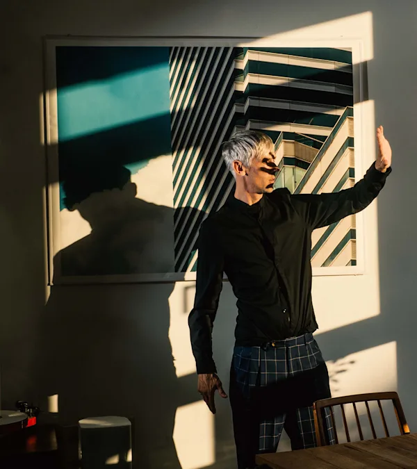A man with silver hair in a black jacket and plaid trousers stands before a framed architectural photo, arm raised, with dramatic shadows.
