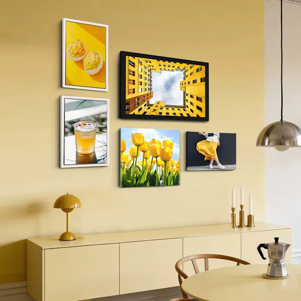 Una disposizione creativa di cinque stampe su tela su una parete giallo chiaro sopra una credenza beige. I soggetti includono architettura, tulipani gialli, una bevanda e nature morte. Le tele sono in parte incorniciate (bianco e nero) e in parte montate senza cornice. Una lampada da tavolo gialla e dei candelabri sulla credenza completano il caldo schema cromatico.