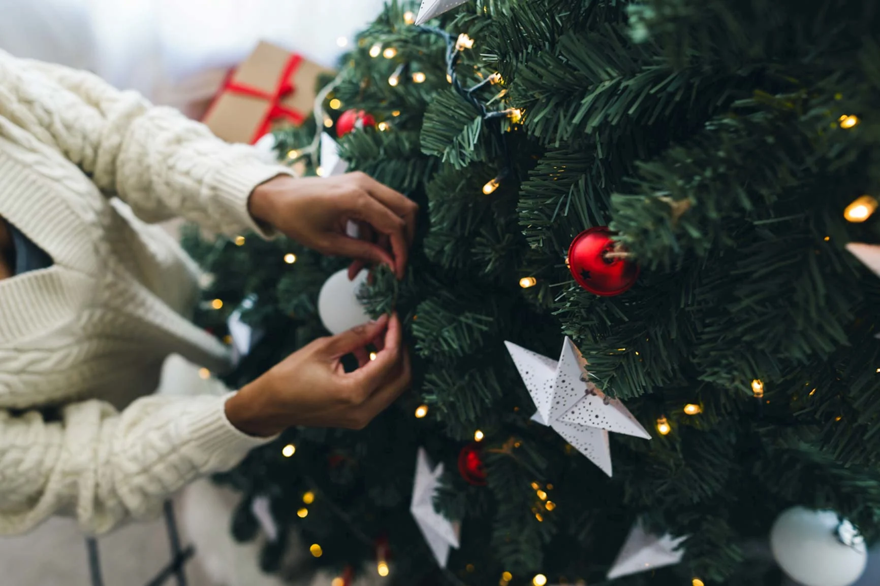 Hände schmücken einen Weihnachtsbaum mit roten Kugeln und weißen Sternen, im Hintergrund funkelnde Lichter und eingepackte Geschenke.