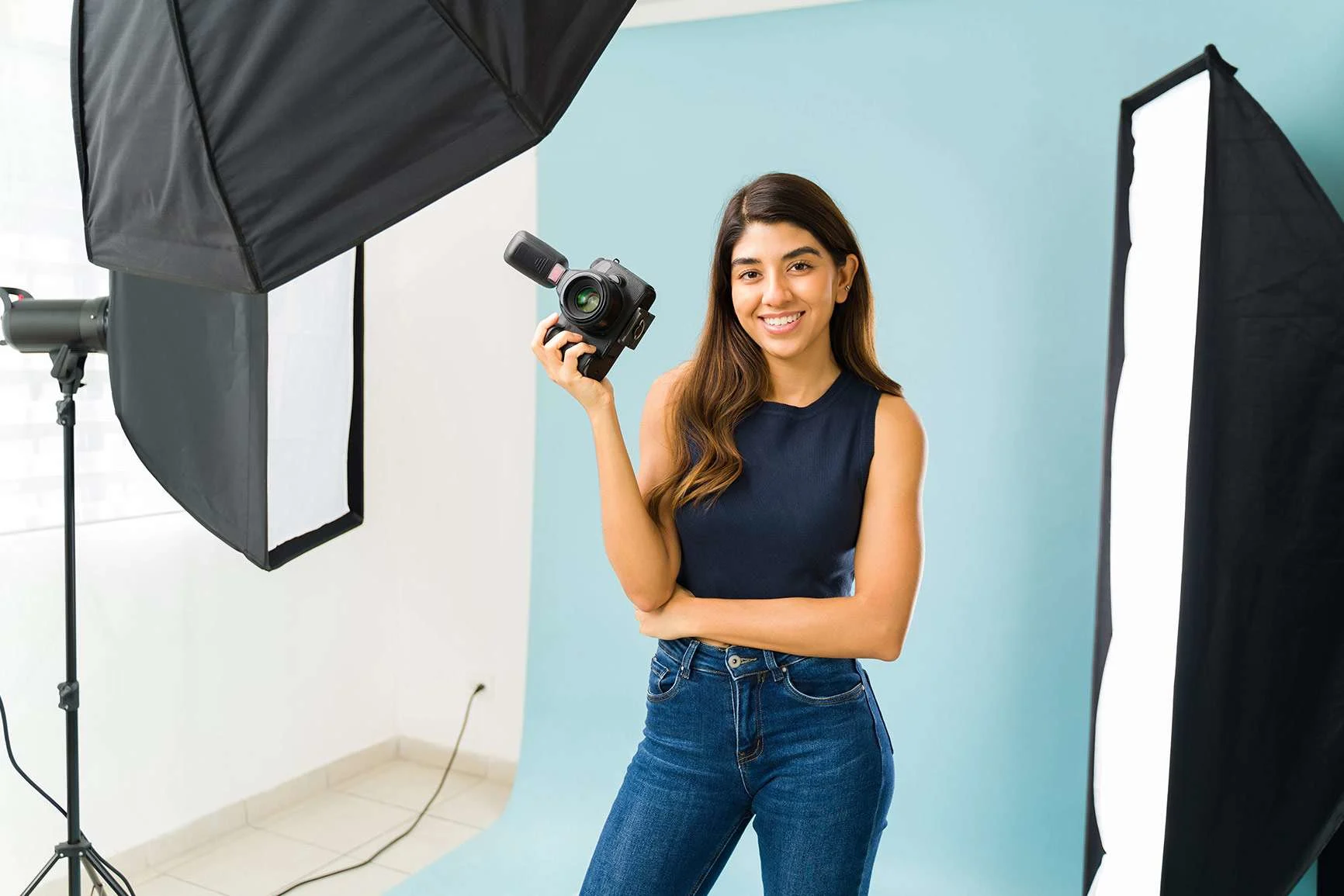 A woman with long hair, wearing a dark top and jeans, is holding a camera in a photo studio with two large softbox lights.