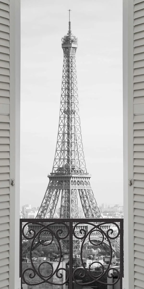 Black and white photograph of the Eiffel Tower seen through an open window with shutters on the side. In the foreground is a wrought-iron railing with curved patterns.