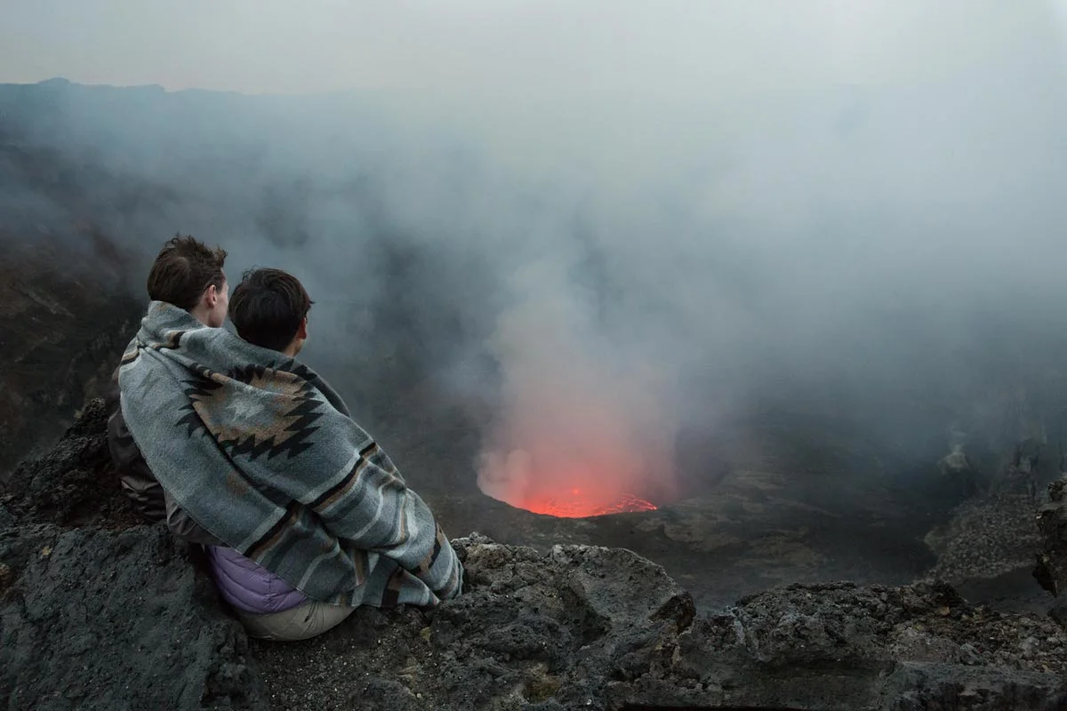 Two people in blankets sit on rocks and look out over a volcano with smoke and glowing lava.