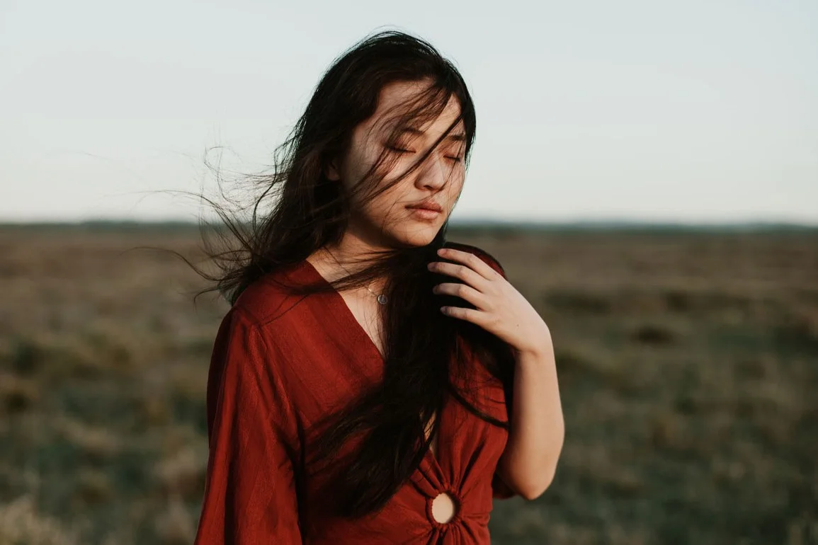 A woman with long dark hair and a red dress is standing outdoors in front of a vast, flat landscape in daylight.