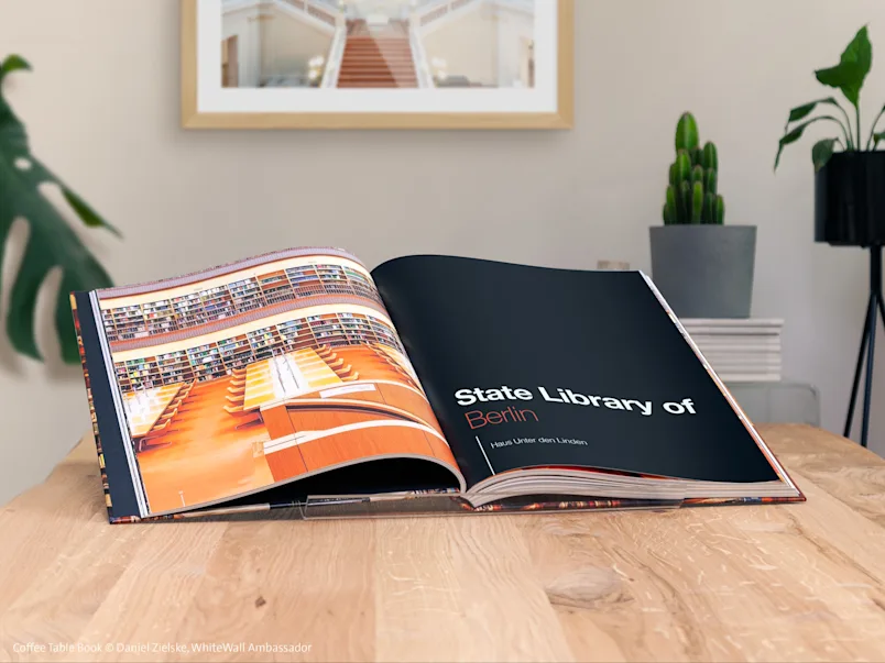 Open photobook on a wooden table, with Berlin State Library photos; plants and photo frame in background.