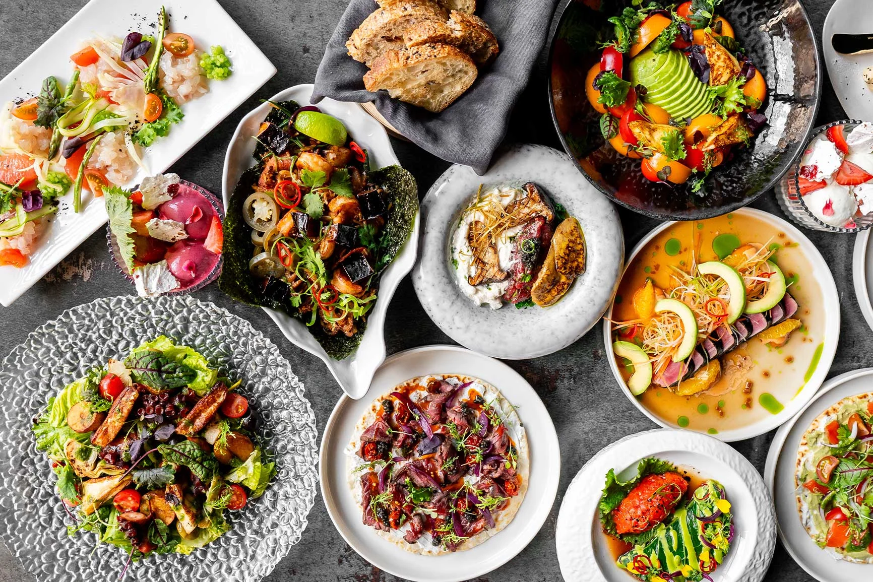 Various artfully arranged dishes featuring vegetables, meat, and bread on plates and bowls set against a gray background.