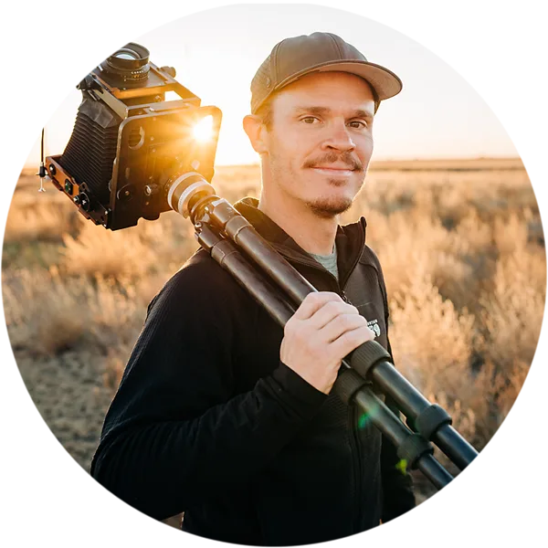 Retrato de Alex Burke con una gorra de béisbol, que lleva al hombro un trípode grande con una cámara, en un paisaje abierto al atardecer.