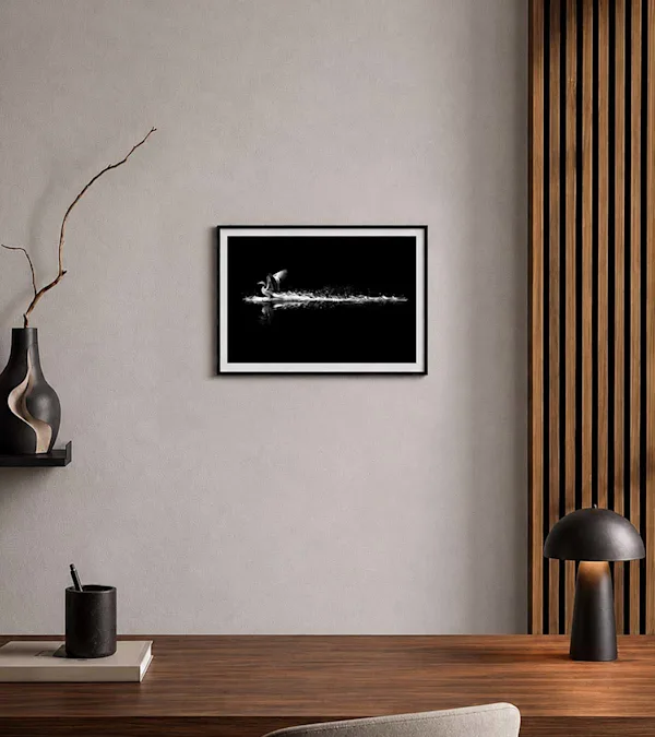 A framed print by Alan Schaller, 'The Landing’ Edition, hanging on a white wall above a wooden desk with a pencil holder and a modern lamp.