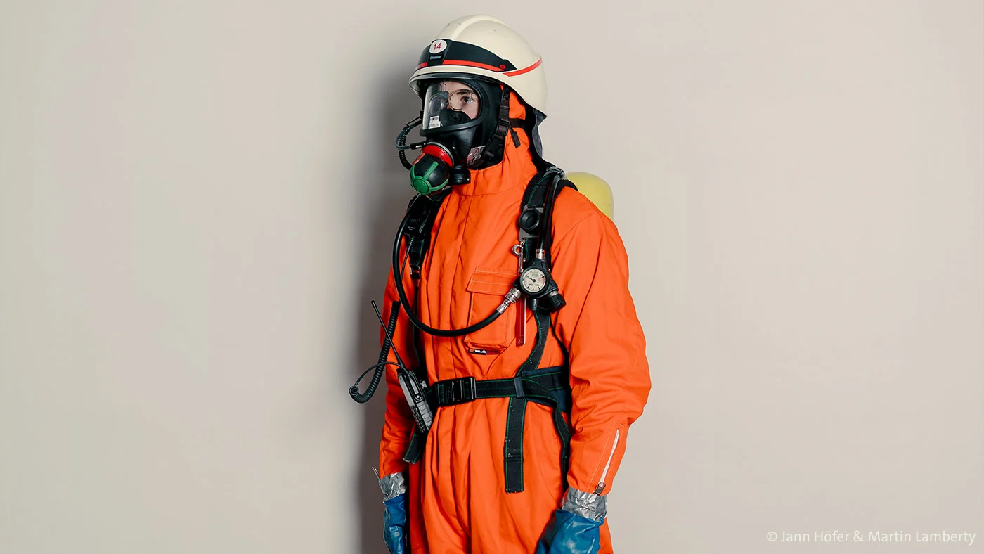 In this striking studio portrait, a figure stands solemnly in full protective gear—an orange hazmat suit, blue gloves, yellow boots, and a respirator mask. The neutral background and clinical lighting draw attention to the uniform’s details, evoking themes of safety, vulnerability, and the human presence behind layers of protection.