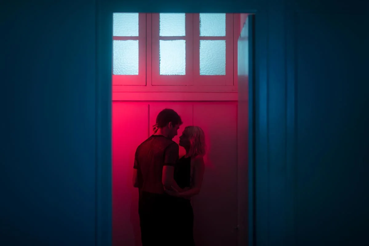 A couple is standing together in a room with red light. The door frame and window above are illuminated in blue.