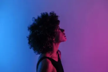 Perfil lateral de una mujer con cabello rizado sobre un fondo azul violeta, mirando hacia arriba, con una camiseta negra.
