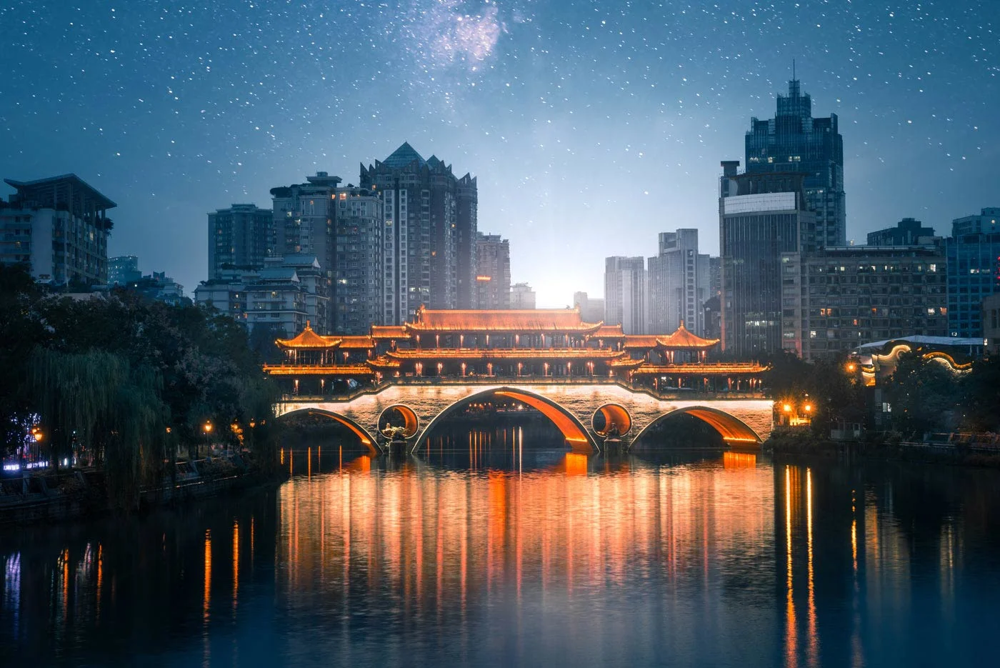 Traditional Chinese bridge with lighting over a river, surrounded by modern high-rise buildings at night.