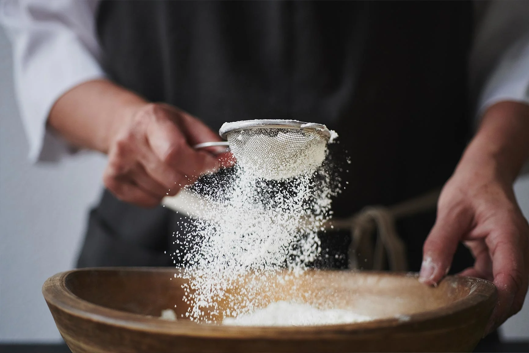 A person sifts flour through a fine sieve into a wooden bowl.