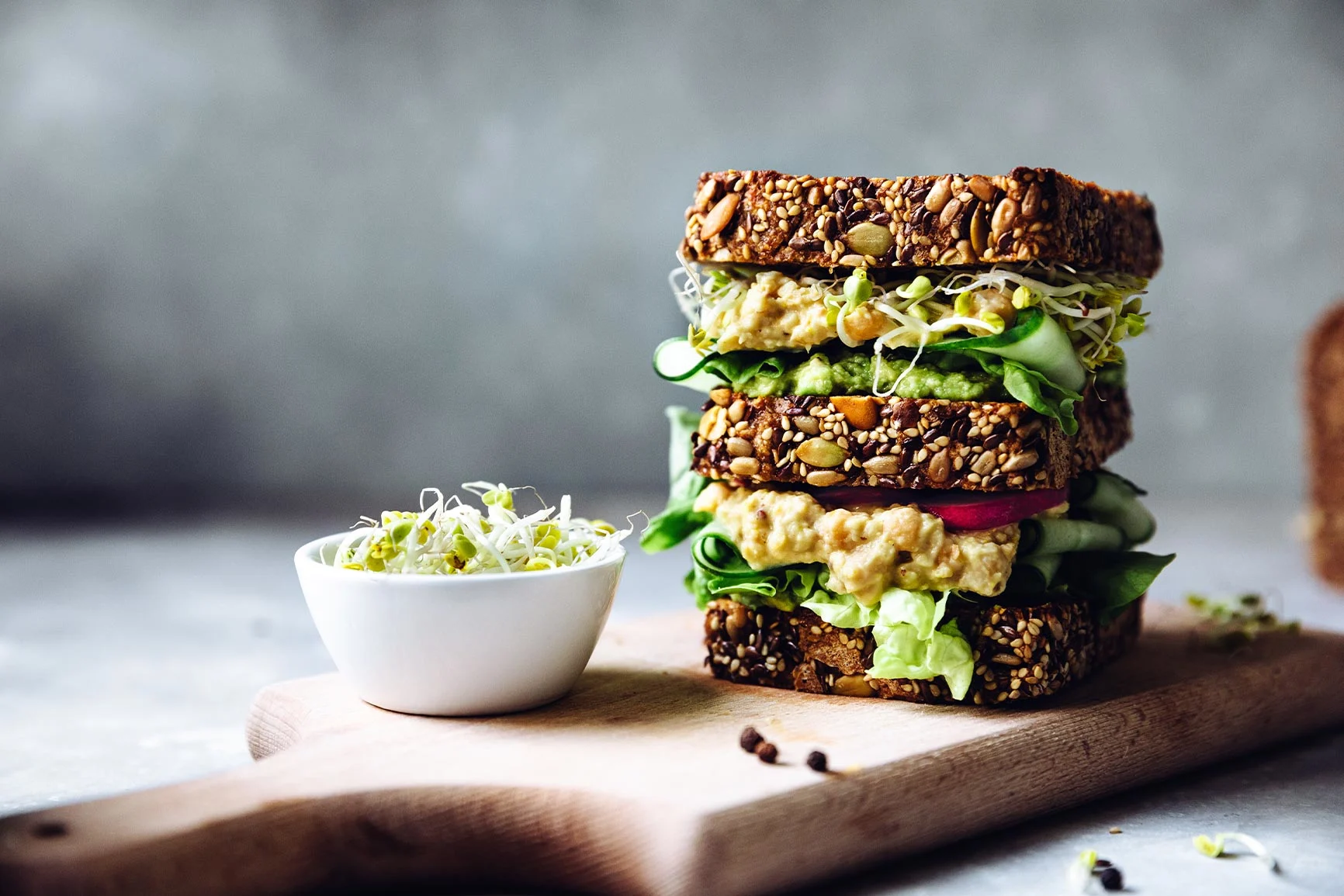 A sandwich made with whole-grain bread, lettuce, cucumber slices, hummus, and sprouts, served on a wooden board next to a small white bowl of sprouts.