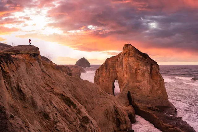 Rock formation on the coast with natural arch, waves, steep cliff, and red sunset sky.