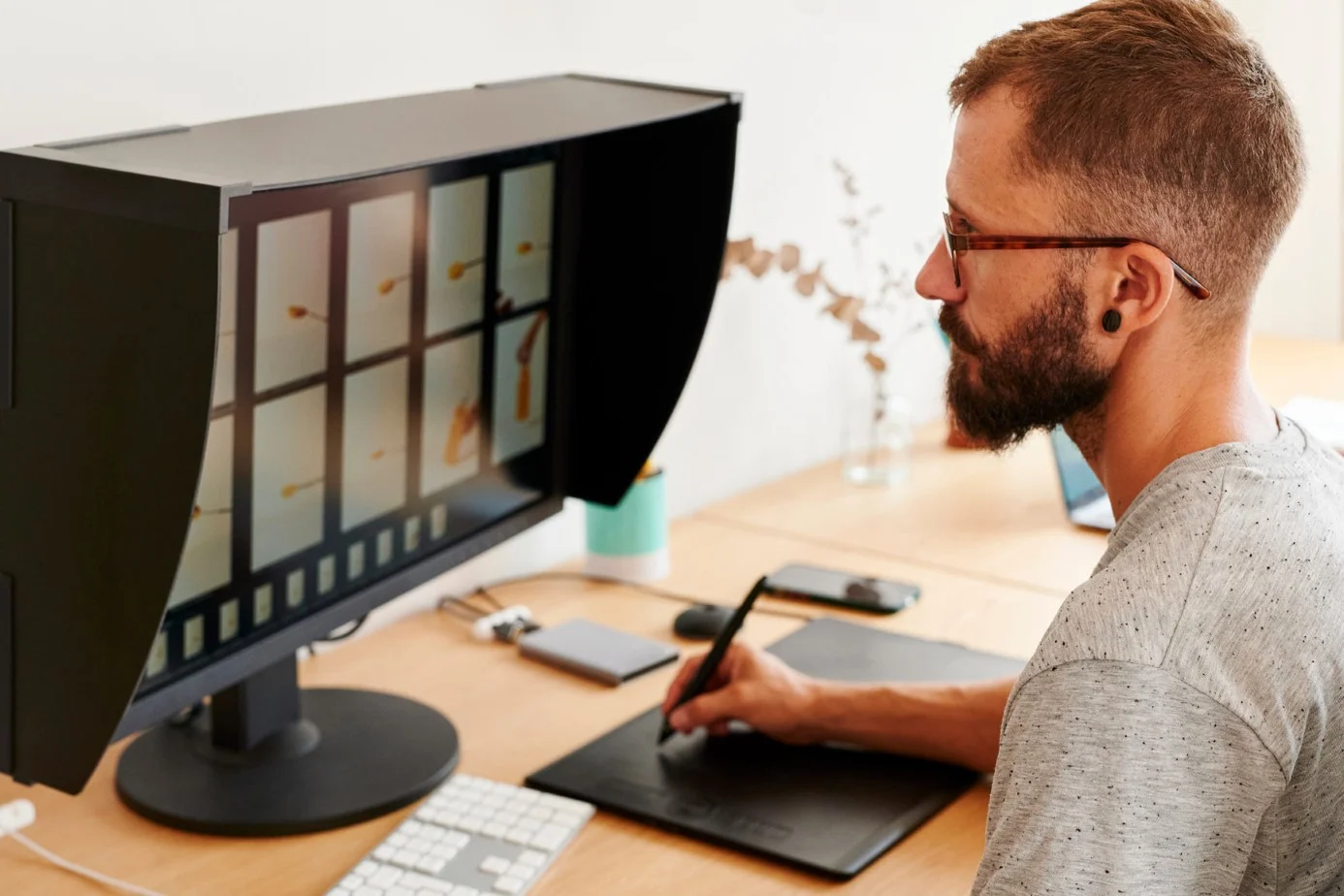 Mann mit Grafiktablett sitzt am Schreibtisch vor einem Monitor mit Lichtschutzhaube und bearbeitet ein Bild.
