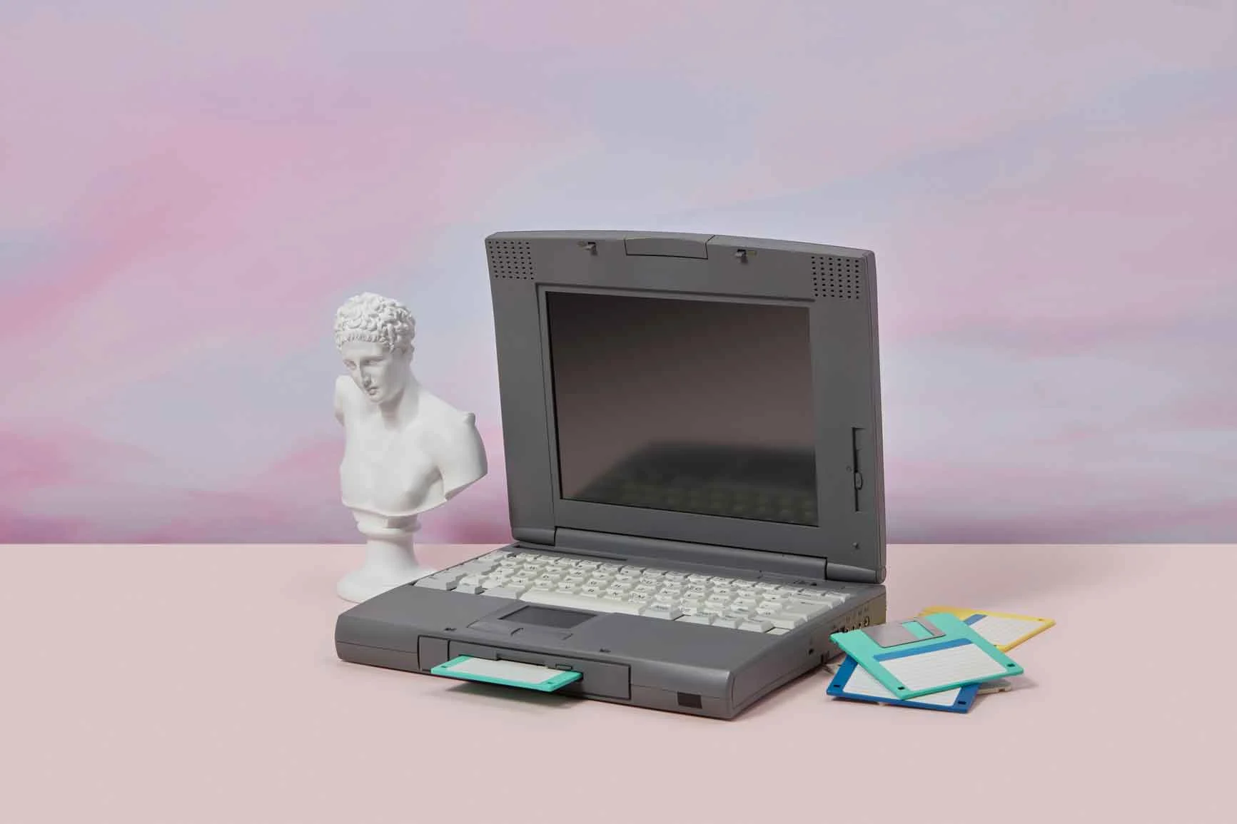 A grey, older laptop stands on a pink background, next to it a white bust and several colorful floppy disks.