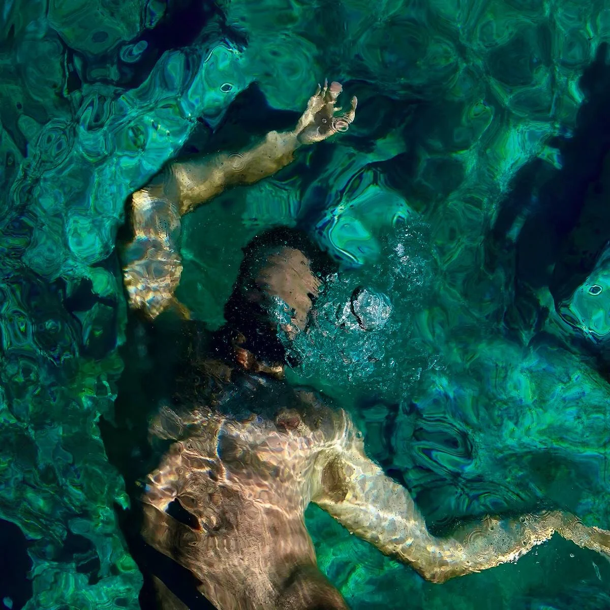 Man swimming underwater with arms outstretched, water surface showing light reflections and shades of green and blue.