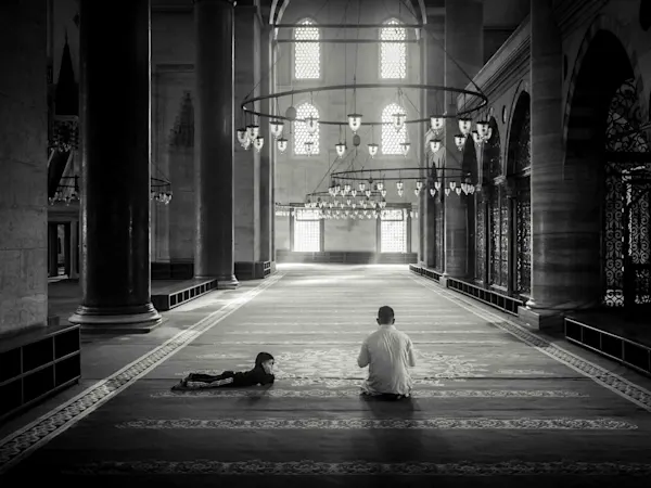 An atmospheric black and white photograph from inside a mosque. In the center, a man sits praying on the carpet while a child lies next to him. The architecture is defined by tall columns, large arched windows in the background, and magnificent circular chandeliers hanging low from the ceiling. The incoming light creates a peaceful, spiritual atmosphere.