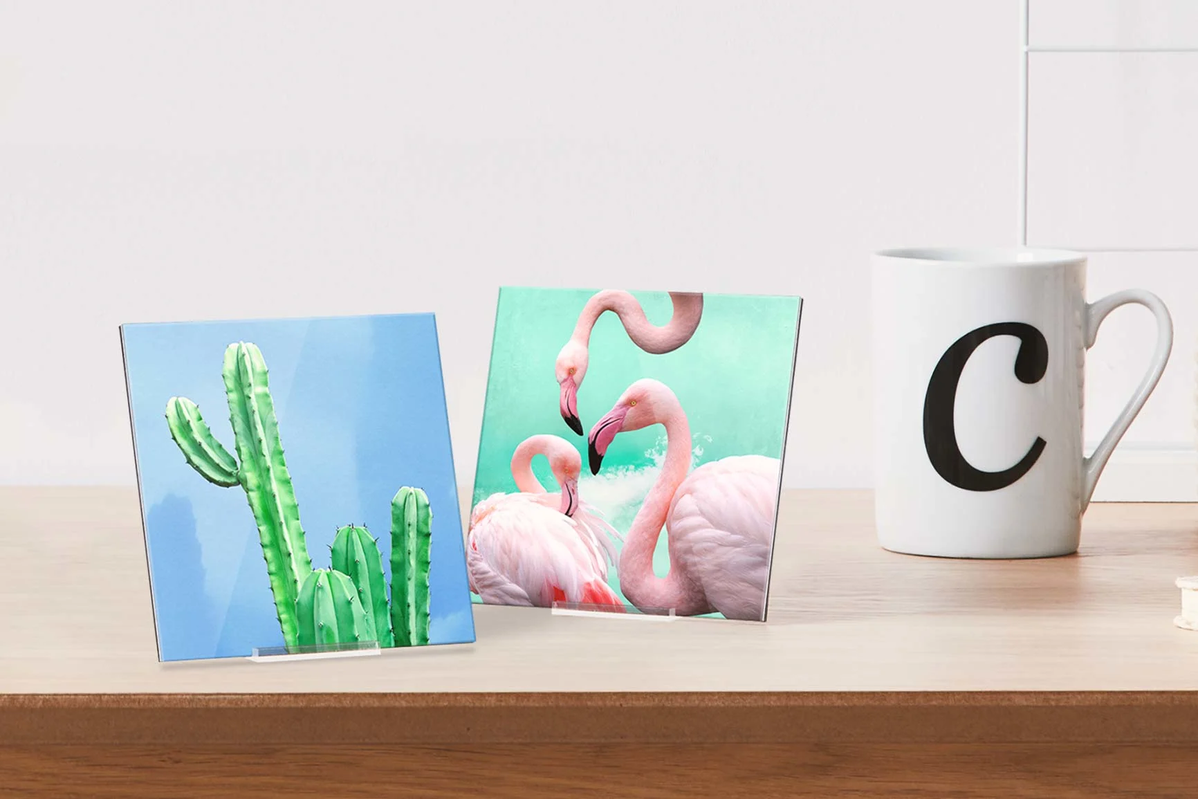 Due espositori fotografici in plexiglass con un motivo ciascuno, a sinistra un cactus, a destra tre fenicotteri, accanto una tazza bianca.