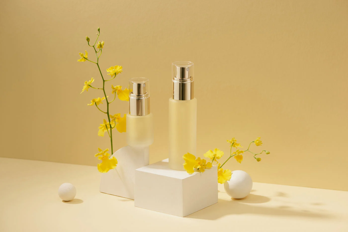 Two transparent pump dispenser bottles containing yellow liquid, surrounded by yellow flowers on a beige background.