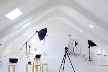 White photo studio with three studio flashes, camera on a tripod, stools, and large background.