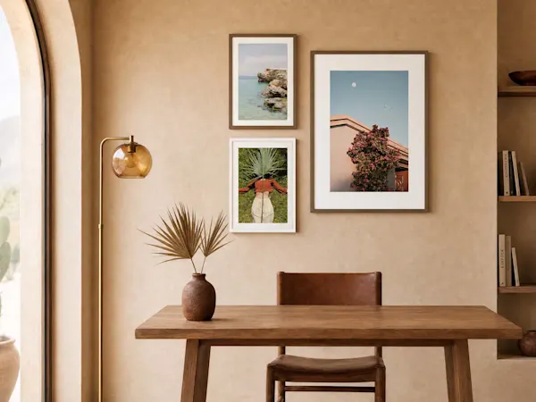 Interior de comedor minimalista con tres impresiones artísticas enmarcadas dispuestas en una pared beige de tono cálido. El marco de madera superior izquierdo muestra una escena costera rocosa con agua turquesa, mientras que el marco de abajo representa a una persona vista desde arriba sosteniendo una gran hoja de palmera sobre su cabeza. A la derecha, un marco vertical más grande exhibe la fachada de un edificio rosa bajo un cielo azul claro con una luna creciente. Debajo de las obras, una mesa de comedor de madera rústica sostiene un jarrón de cerámica con hojas de palmera secas frente a una silla de cuero a juego.