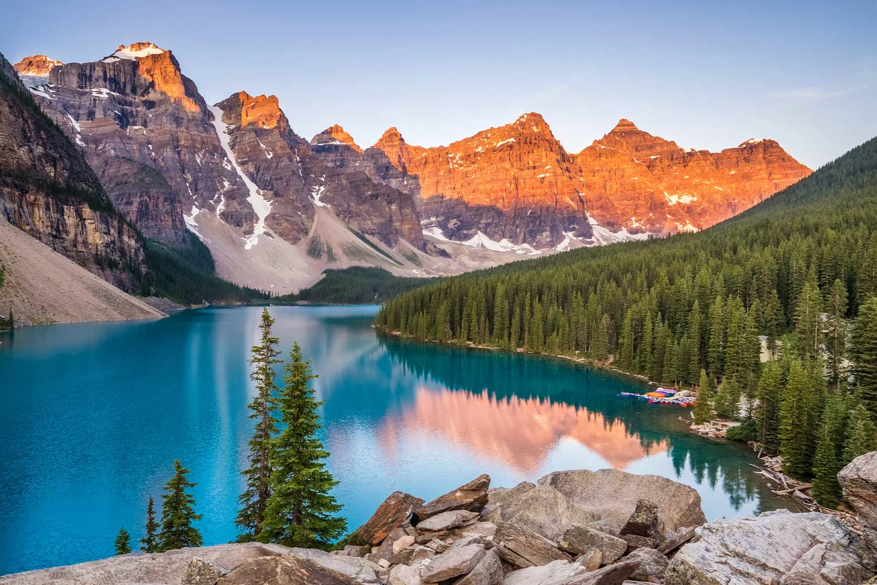 A mountain lake with a clear surface, surrounded by coniferous forest and rocks, with glowing mountain peaks in the background bathed in the evening light.