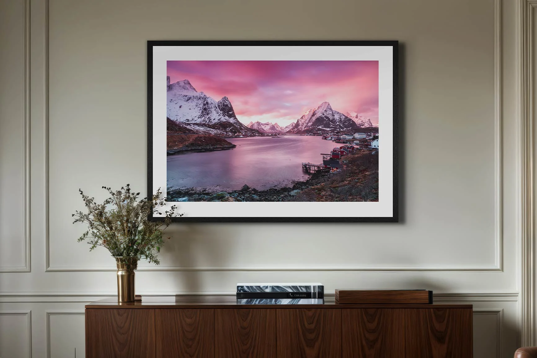 A framed landscape photo featuring mountains and water under a pink sky, hanging above a sideboard with a vase and books.